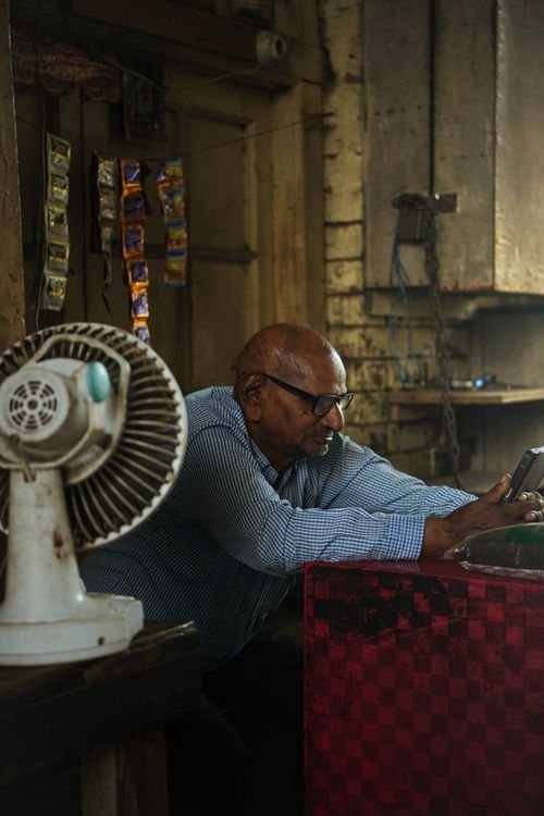 A shopkeeper in a small shop leaning on a counter and looking at a phone, with a fan beside him and shop goods behind him