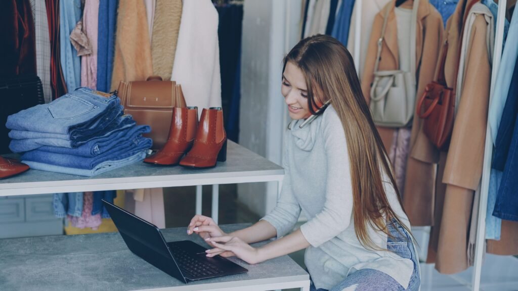 A young woman in a fashion boutique, talking on a phone while working on a laptop among clothing