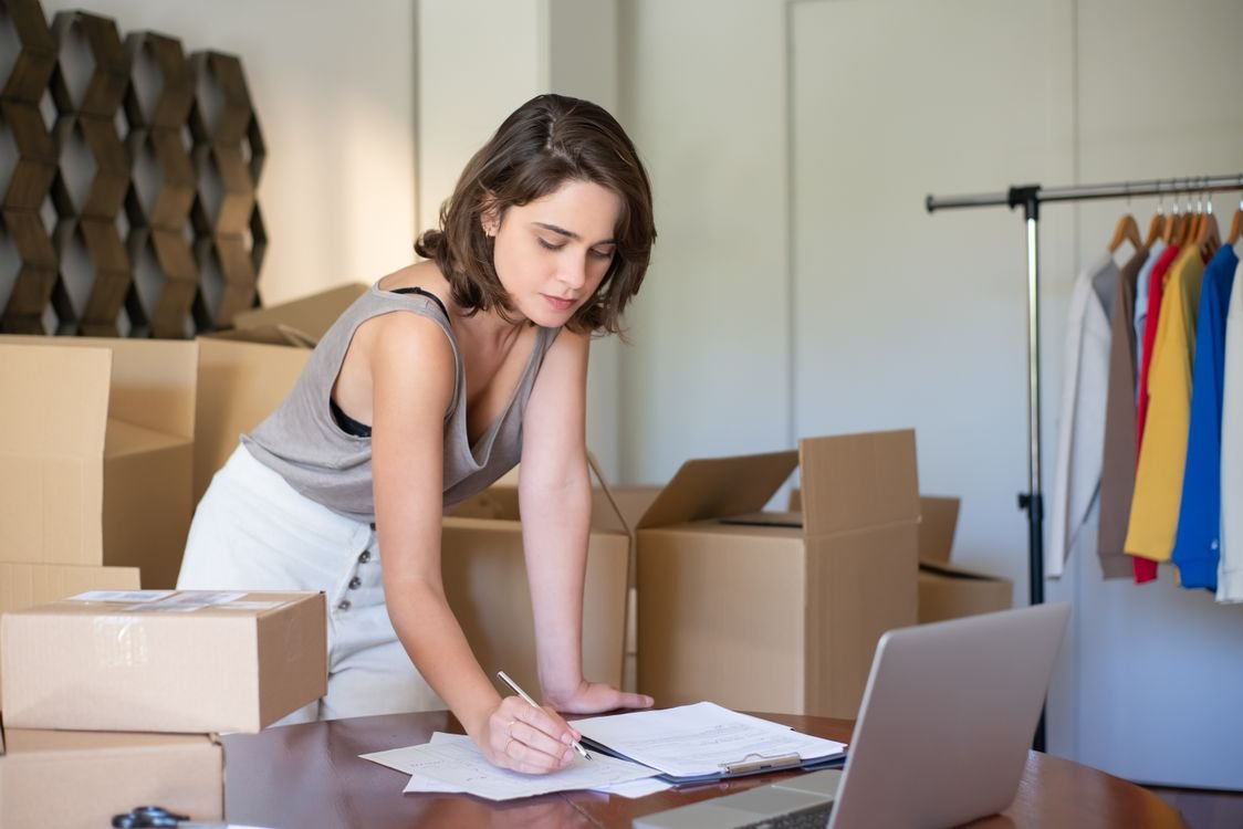 A woman managing an online business from a home office, standing among cardboard boxes and writing at a desk with a laptop