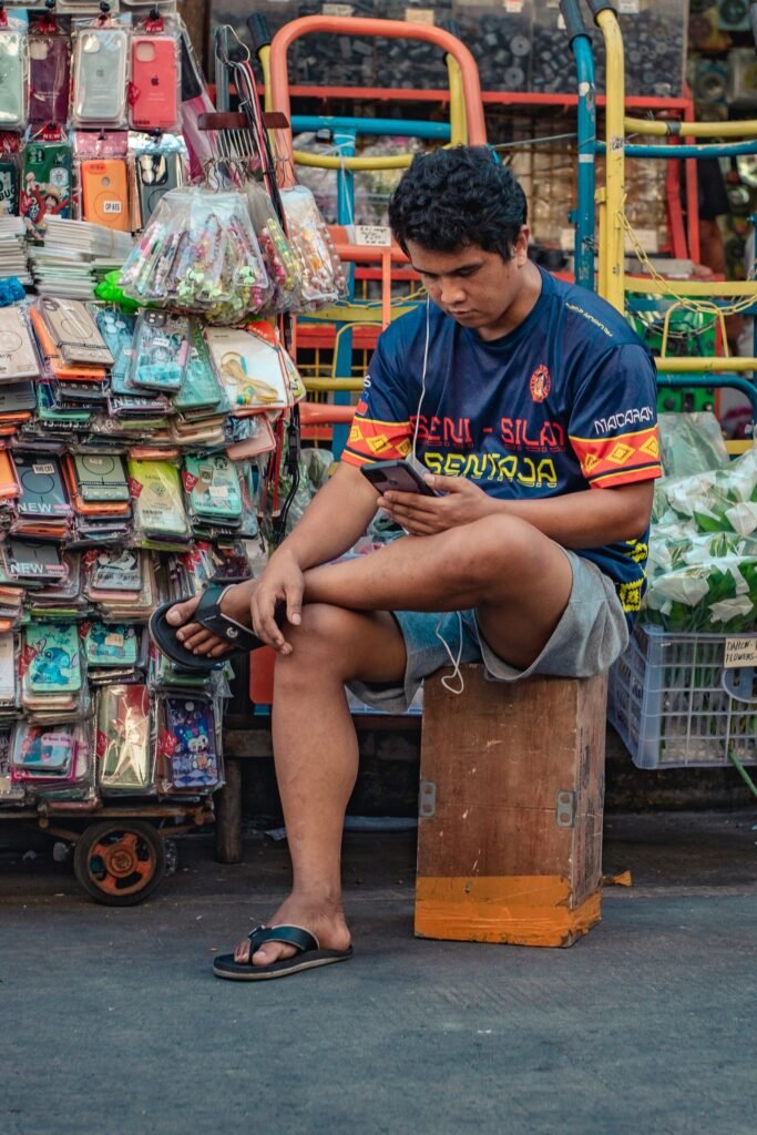 A street vendor sits among colourful merchandise in a busy outdoor market, focused on his smartphone.