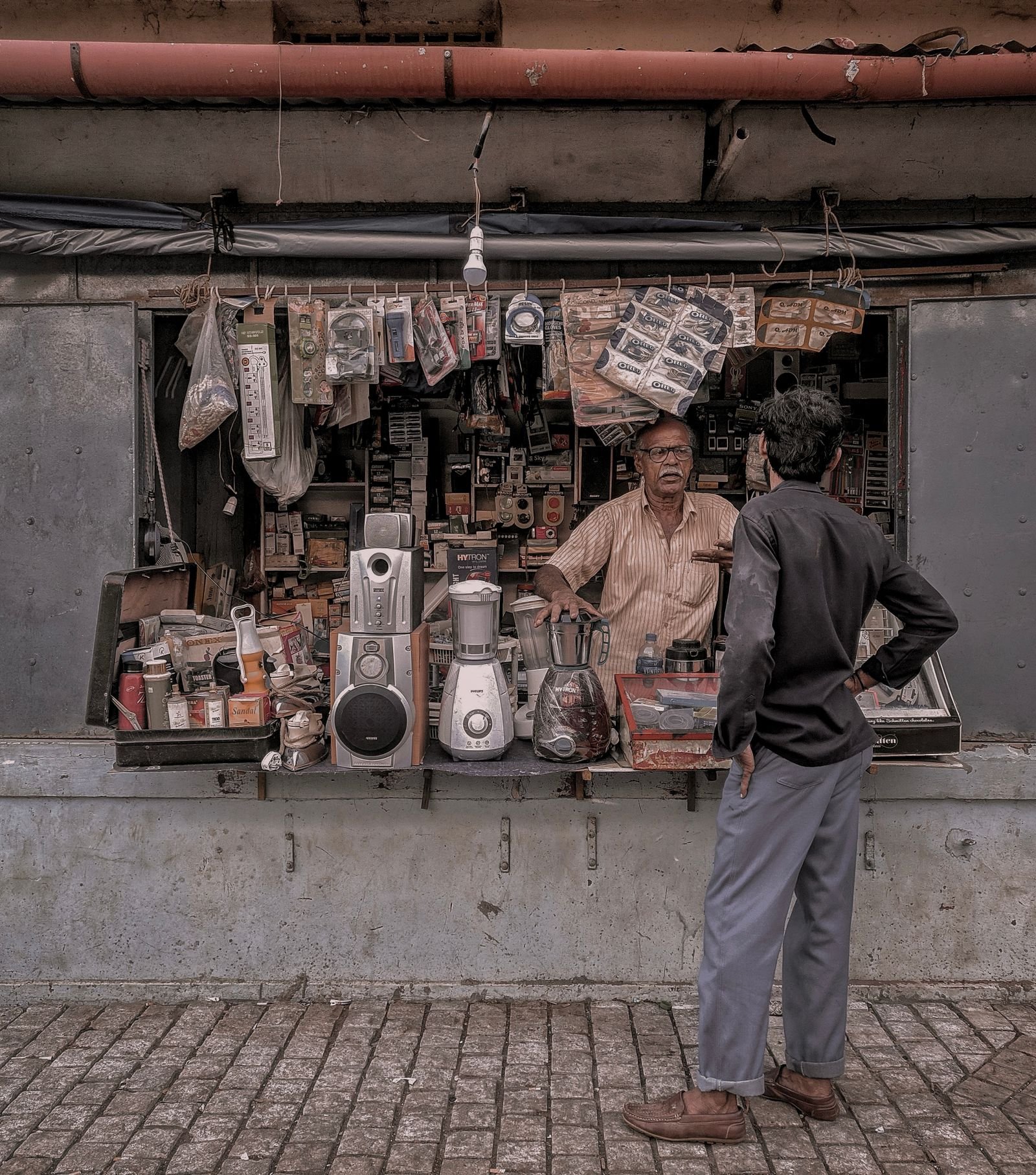A vendor talks with a customer at a small electronics stall in a local marketplace.