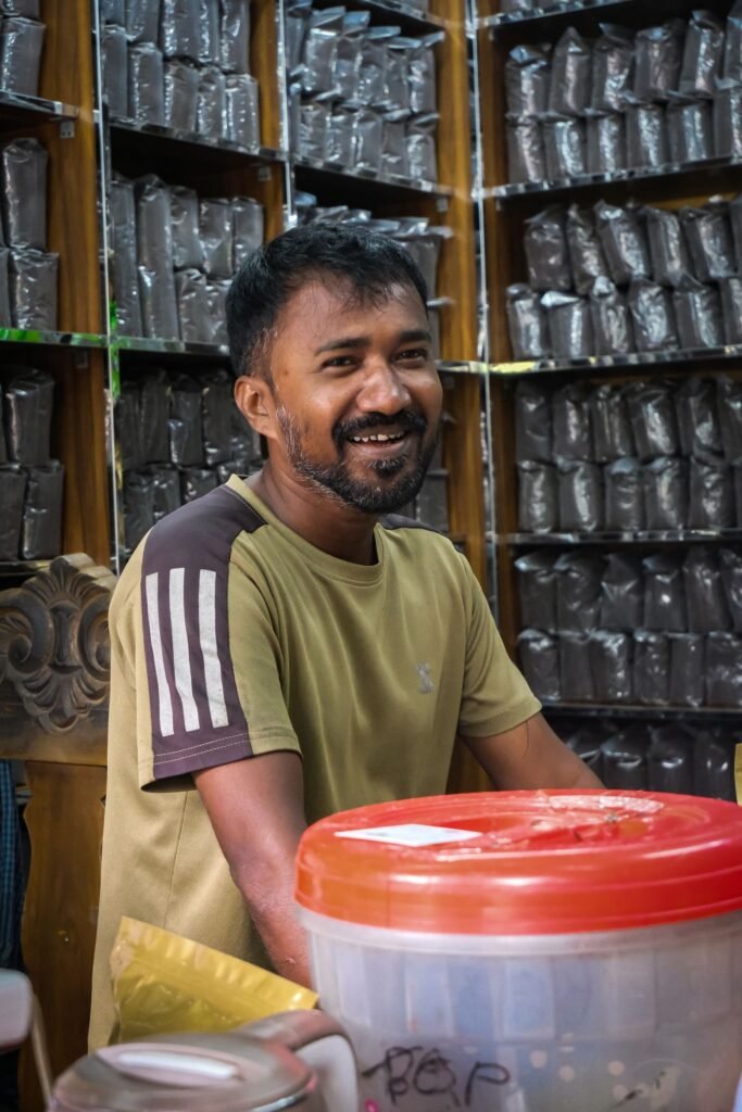 A smiling shopkeeper stands in his tea store among shelves of packaged products.