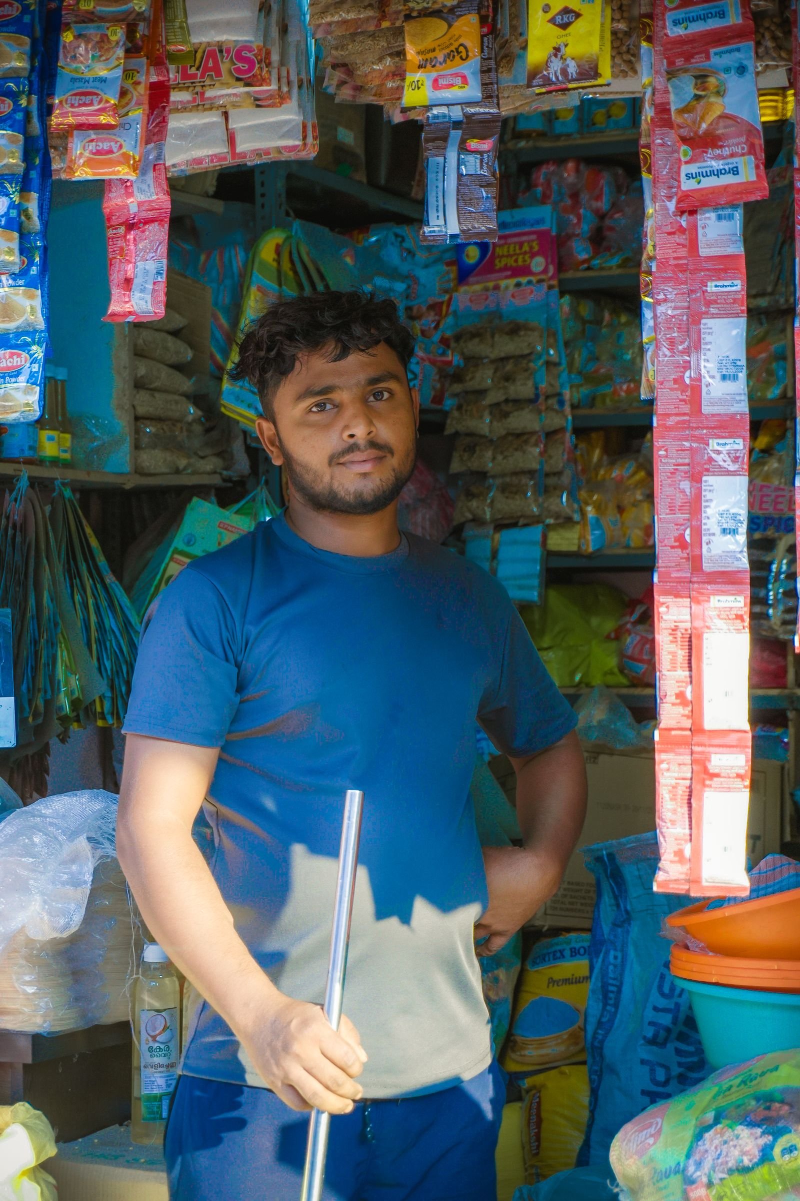 A shopkeeper stands among hanging snacks and stocked goods in a local grocery store.